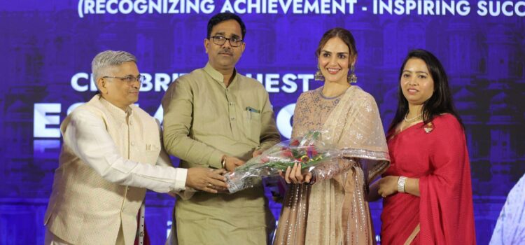 Four individuals on stage, with one man handing a bouquet of flowers to a Esha Deol in a beige traditional outfit, against a backdrop displaying "Excellency Iconic Awards" and "Recognizing Achievement - Inspiring Success.